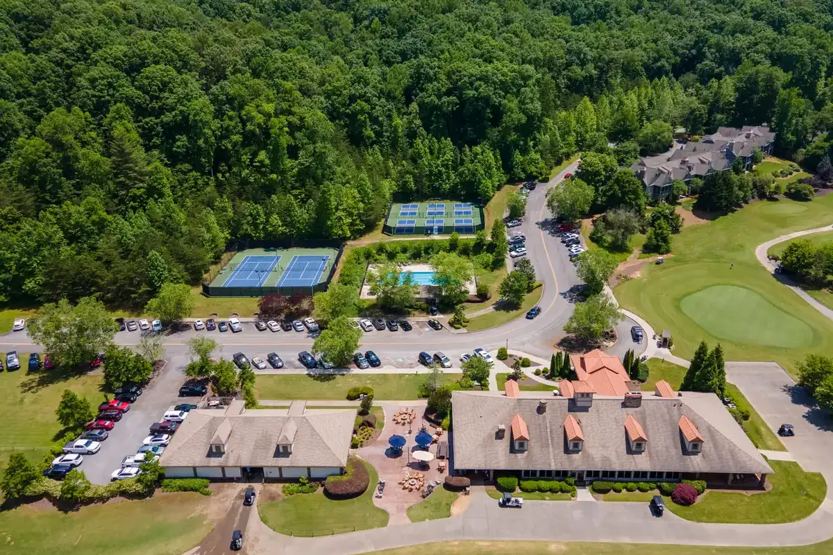 Aerial view of the Achasta community amenity campus on a sunny day: the dormered clubhouse and patio, tennis and pickleball courts, outdoor swimming pool, manicured golf green with cart paths, parked cars, and condo buildings tucked into dense North Georgia forest.