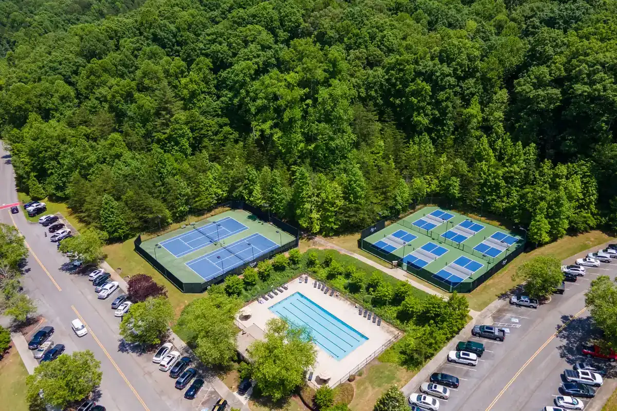 Aerial view of tennis and pickleball courts and a fenced lap pool set into dense green forest, with parking along a tree-lined road