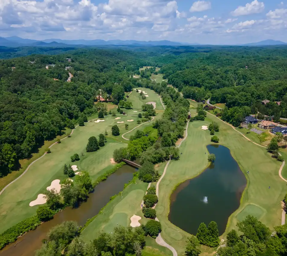 Aerial drone view of the Achasta golf course: green fairways with sand bunkers winding through dense forest, a large pond with a fountain, a river with a wooden bridge, homes along the fairway edge, and layered Blue Ridge mountains under a partly cloudy sky.