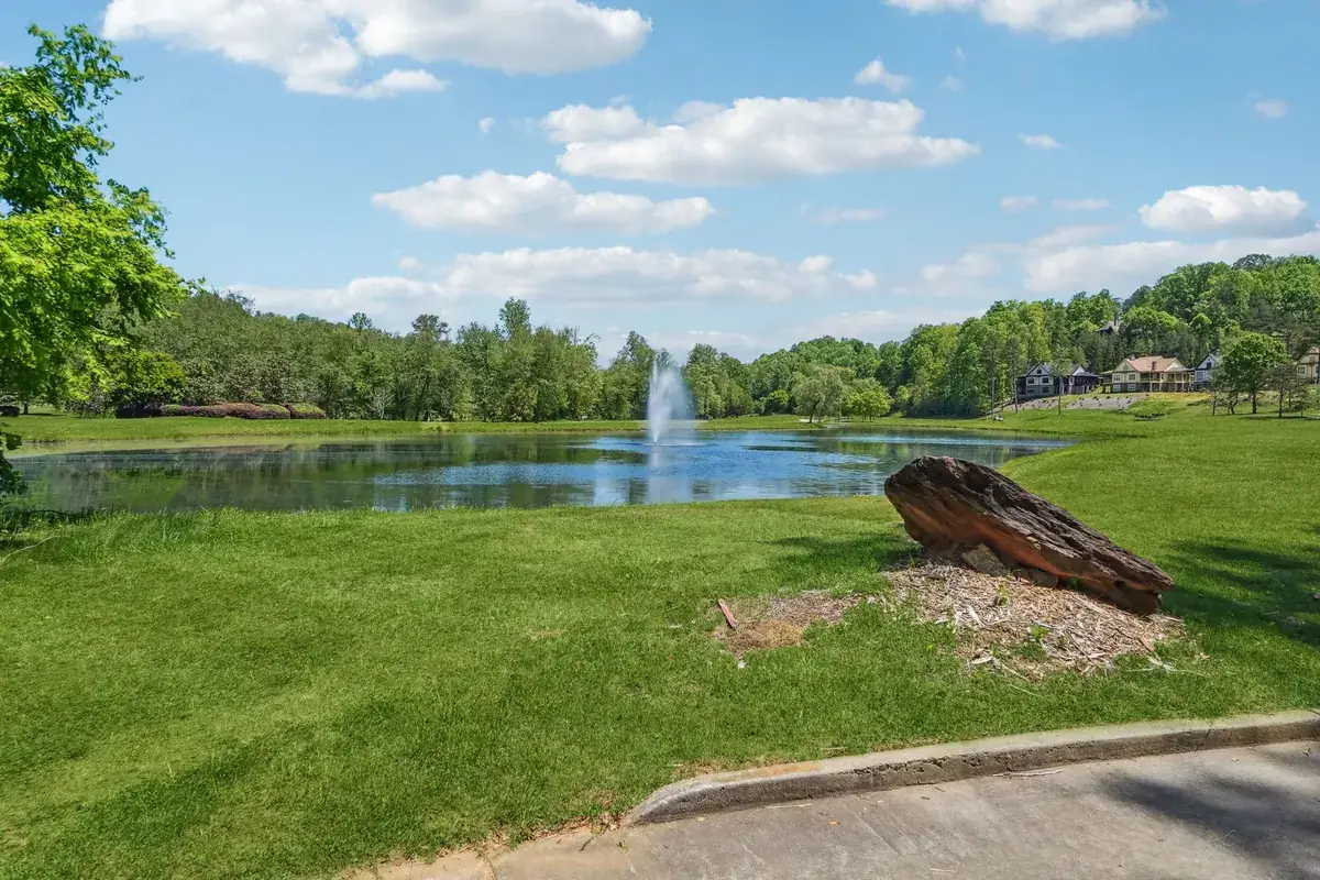 Pond with a fountain on a wide green lawn, trees along the far bank and homes on the wooded slope beyond