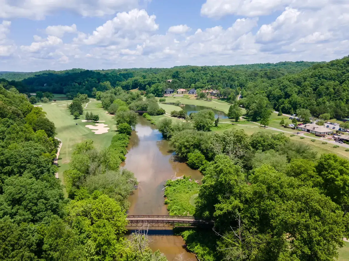 Aerial view of the Chestatee River running through the Achasta golf community in Dahlonega, Georgia, with a footbridge in the foreground, fairways and sand bunkers on the left bank, and homes on the wooded ridge beyond.